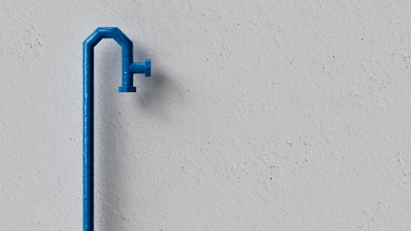 A minimalist scene featuring a blue water pipe against a plain, textured gray wall. The pipe has a slightly curved shape with a spout extending outward.