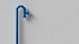 A minimalist scene featuring a blue water pipe against a plain, textured gray wall. The pipe has a slightly curved shape with a spout extending outward.