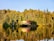 A small cabin with a red roof is nestled among dense forests of tall green and golden trees, situated on the edge of a calm lake. The clear water reflects the vibrant colors of the trees and the cabin, creating a mirror-like effect.