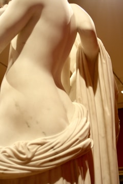 A close-up of a marble sculpture depicting a partially draped human figure with detailed carving. The smooth curves and folds of the material showcase the craftsmanship and evoke a classical aesthetic.