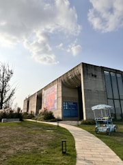 A modern concrete building with large windows and a prominent billboard. The structure is surrounded by green grass and a winding stone path leading up to it. In the foreground, there is a blue and white striped pedal cart. The sky is partly cloudy, with sunlight breaking through.