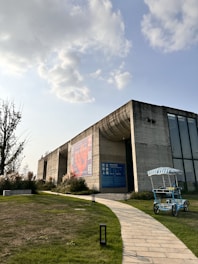 A modern concrete building with large windows and a prominent billboard. The structure is surrounded by green grass and a winding stone path leading up to it. In the foreground, there is a blue and white striped pedal cart. The sky is partly cloudy, with sunlight breaking through.