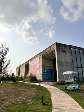 A modern concrete building with large windows and a prominent billboard. The structure is surrounded by green grass and a winding stone path leading up to it. In the foreground, there is a blue and white striped pedal cart. The sky is partly cloudy, with sunlight breaking through.