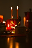 A cozy, warm ambiance is created by a Halloween-themed setup with lit candles. A jack-o'-lantern with glowing eyes and mouth stands prominently in the background. A coffee mug with an inscription sits among other items, possibly a slice of cake and a pot. The dim lighting and reflections add to the intimate atmosphere.