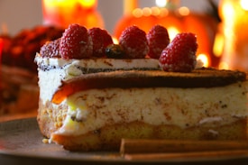 A close-up of a slice of cheesecake topped with fresh raspberries. The dessert sits on a plate, surrounded by warm, soft lighting that suggests a cozy atmosphere. Cinnamon sticks can be seen on the side, adding a rustic touch.