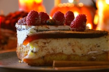 A close-up of a slice of cheesecake topped with fresh raspberries. The dessert sits on a plate, surrounded by warm, soft lighting that suggests a cozy atmosphere. Cinnamon sticks can be seen on the side, adding a rustic touch.