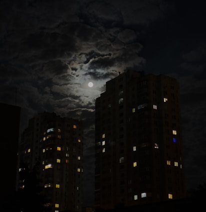 Two tall apartment buildings with illuminated windows stand against a dark, cloudy night sky. The full moon shines brightly, casting a mystical glow around the clouds.