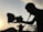 A silhouette of a person operating a film camera is prominently featured against a backdrop of a sunset. The sky is softly illuminated, with power lines visible in the distance. Shadows cast intricate shapes along the contours of the person and equipment.