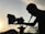 A silhouette of a person operating a film camera is prominently featured against a backdrop of a sunset. The sky is softly illuminated, with power lines visible in the distance. Shadows cast intricate shapes along the contours of the person and equipment.