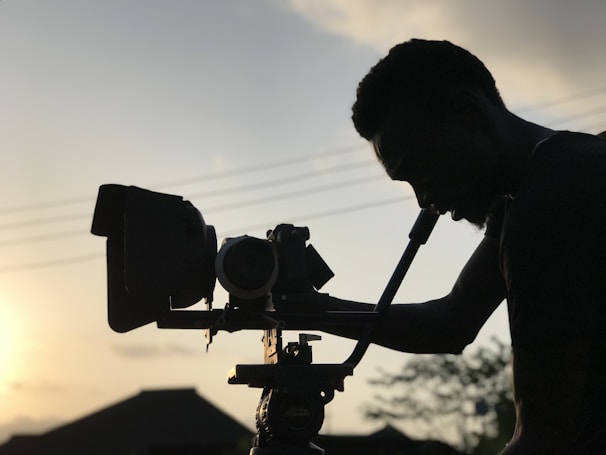 A silhouette of a person operating a film camera is prominently featured against a backdrop of a sunset. The sky is softly illuminated, with power lines visible in the distance. Shadows cast intricate shapes along the contours of the person and equipment.