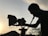 A silhouette of a person operating a film camera is prominently featured against a backdrop of a sunset. The sky is softly illuminated, with power lines visible in the distance. Shadows cast intricate shapes along the contours of the person and equipment.