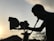 A silhouette of a person operating a film camera is prominently featured against a backdrop of a sunset. The sky is softly illuminated, with power lines visible in the distance. Shadows cast intricate shapes along the contours of the person and equipment.