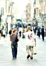 A busy urban street scene with people walking along a pedestrian area. The focus is on two individuals from behind, one wearing a graphic t-shirt and holding a shopping bag, while the other is dressed in casual jeans. The street is lined with tall buildings, and the atmosphere is lively with several people in the background.