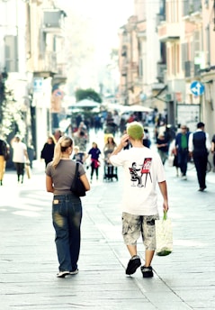 A busy urban street scene with people walking along a pedestrian area. The focus is on two individuals from behind, one wearing a graphic t-shirt and holding a shopping bag, while the other is dressed in casual jeans. The street is lined with tall buildings, and the atmosphere is lively with several people in the background.