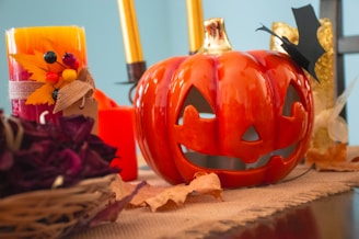 A Halloween-themed scene featuring a ceramic pumpkin with a carved jack-o'-lantern face on a table. Beside it, there are decorative candles with autumnal embellishments like leaves and berries. Golden decorative elements and dried autumn leaves are placed around, contributing to the seasonal theme.