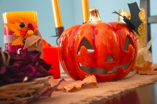 A Halloween-themed scene featuring a ceramic pumpkin with a carved jack-o'-lantern face on a table. Beside it, there are decorative candles with autumnal embellishments like leaves and berries. Golden decorative elements and dried autumn leaves are placed around, contributing to the seasonal theme.