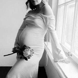 A pregnant woman wearing an off-shoulder dress leans against a windowsill, holding a large flower by her side. The setting is softly lit, highlighting her belly and creating a serene atmosphere.