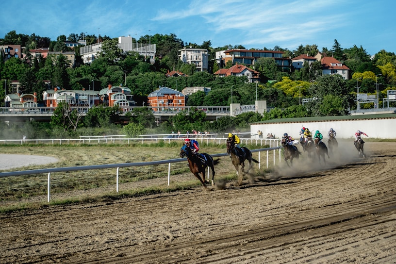 A group of horses with jockeys racing on a dirt track, surrounded by lush greenery and residential buildings in the background. Dust is kicking up from the horses' hooves as they round a bend.