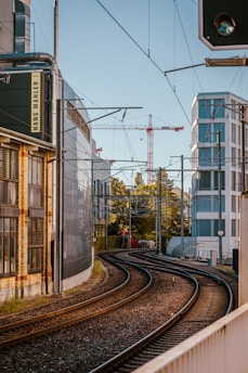 Curved railroad tracks run through an urban area, bordered by modern buildings with glass facades and industrial structures. Overhead power lines and a distant red crane are visible against a clear sky, with trees providing greenery in the background.