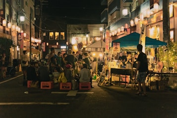 A bustling night market with a group of people gathered around food stalls. Lanterns hang above, illuminating the scene with a warm glow. Vendors are serving food under a blue canopy, and some people are seated on crates enjoying the atmosphere. The street is lined with illuminated buildings, adding to the lively ambiance.