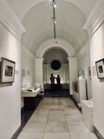 A corridor in a museum features a high arched ceiling and walls adorned with framed artwork. Two people are standing at the far end, observing items housed in glass display cases. The lighting is soft and creates a contemplative atmosphere.