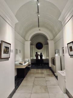 A corridor in a museum features a high arched ceiling and walls adorned with framed artwork. Two people are standing at the far end, observing items housed in glass display cases. The lighting is soft and creates a contemplative atmosphere.