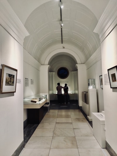A corridor in a museum features a high arched ceiling and walls adorned with framed artwork. Two people are standing at the far end, observing items housed in glass display cases. The lighting is soft and creates a contemplative atmosphere.