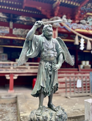 A bronze statue of a warrior with wings, standing on a rock pedestal. The warrior is dressed in traditional attire, with one hand holding a sword raised above the head. The background shows a traditional architectural structure with intricate designs and a wooden fence.