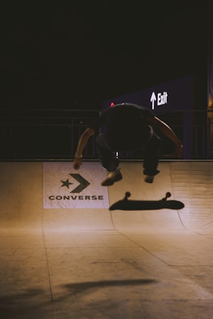 A skateboarder in mid-air performing a trick in a dimly lit skatepark. The silhouette of the person is blurred against the bright background, which features a large Converse logo on the ramp wall and an illuminated exit sign in the distance. The atmosphere is moody with high contrast between shadow and light.