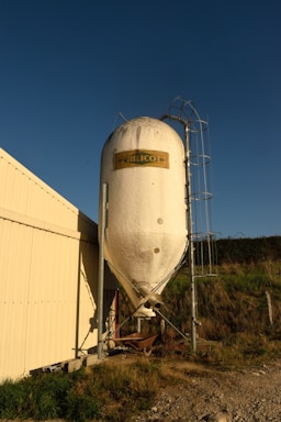 A large vertical storage silo tank stands next to a light-colored industrial building. The tank is cylindrical and white, with a slightly conical bottom. An emblem with the name 'HILICO' is affixed to the upper part of the tank. A metal ladder attached to the rack is visible on the right side, reaching up the tank. The area is surrounded by grass and a dirt path, with clear blue skies in the background.
