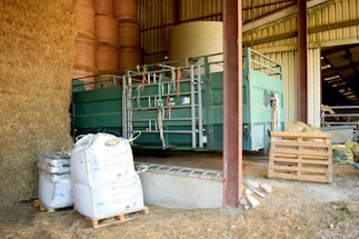 Inside a barn, there are several large bales of hay stacked high. A green piece of farm machinery, possibly used for animal handling, stands nearby. Next to it are multiple large bags of feed on pallets, and some scattered wooden pallets on the ground. The interior of the barn is partially visible, with a roof and metal beams.