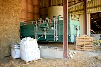 Inside a barn, there are several large bales of hay stacked high. A green piece of farm machinery, possibly used for animal handling, stands nearby. Next to it are multiple large bags of feed on pallets, and some scattered wooden pallets on the ground. The interior of the barn is partially visible, with a roof and metal beams.