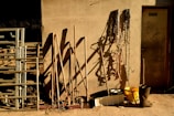 A rustic scene with various farming tools leaning against a textured wall. Shadows cast by the tools are prominent on the wall. Metal pipes or railing are stacked to the left, and coiled ropes hang on the right. A yellow bucket and a pair of black boots are placed on the ground.