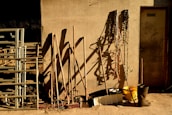 A rustic scene with various farming tools leaning against a textured wall. Shadows cast by the tools are prominent on the wall. Metal pipes or railing are stacked to the left, and coiled ropes hang on the right. A yellow bucket and a pair of black boots are placed on the ground.