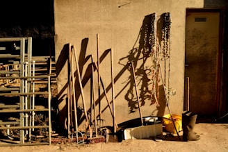 A rustic scene with various farming tools leaning against a textured wall. Shadows cast by the tools are prominent on the wall. Metal pipes or railing are stacked to the left, and coiled ropes hang on the right. A yellow bucket and a pair of black boots are placed on the ground.