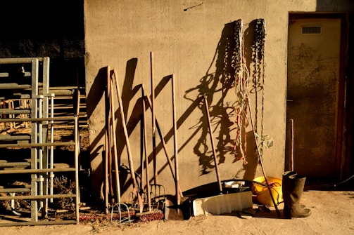 A rustic scene with various farming tools leaning against a textured wall. Shadows cast by the tools are prominent on the wall. Metal pipes or railing are stacked to the left, and coiled ropes hang on the right. A yellow bucket and a pair of black boots are placed on the ground.