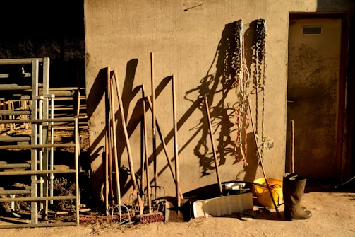 A rustic scene with various farming tools leaning against a textured wall. Shadows cast by the tools are prominent on the wall. Metal pipes or railing are stacked to the left, and coiled ropes hang on the right. A yellow bucket and a pair of black boots are placed on the ground.