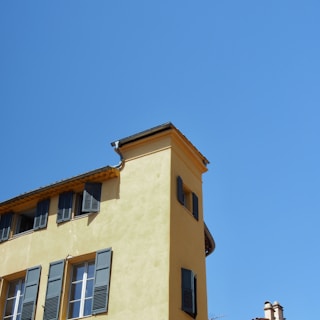 A building with a yellow facade and blue shutters set against a clear blue sky. The structure features multiple windows and a roof with visible gutters.