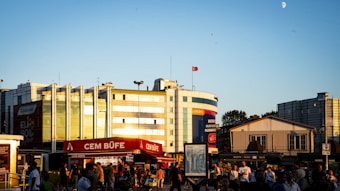A busy urban setting with several people walking in front of commercial buildings under the evening sky. A red storefront labeled 'Cem B&uuml;fe' is prominent in the foreground, with modern architectural structures and billboards in the background. A few flags are visible on top of the buildings, and the sky is clear with a visible moon.