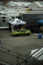 A vivid green sports car is parked in a lot surrounded by white tents and trailers. The car is positioned under a canopy with the words 'Brumos.com' visible. The area appears to be part of a racetrack or car event setup. The ground is cracked pavement, and there are blue barrels and a checkered-patterned bin nearby.