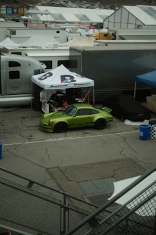 A vivid green sports car is parked in a lot surrounded by white tents and trailers. The car is positioned under a canopy with the words 'Brumos.com' visible. The area appears to be part of a racetrack or car event setup. The ground is cracked pavement, and there are blue barrels and a checkered-patterned bin nearby.