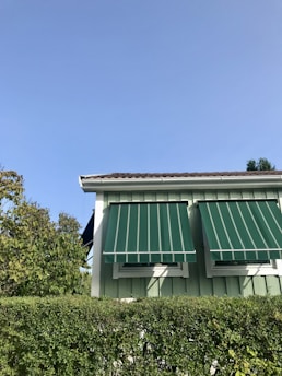 A suburban house with light green wooden siding features two windows with green and white striped awnings. In front of the house is a well-trimmed hedge, with trees partially visible on the side. The sky above is clear and bright, suggesting a sunny day.