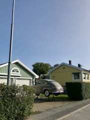 A classic car is elevated on jacks in front of two suburban houses, one painted green and the other yellow, both with enclosed garages. A streetlight and neatly trimmed bushes line the sidewalk, and trees are visible in the background under a clear blue sky.