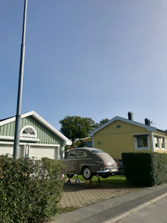 A classic car is elevated on jacks in front of two suburban houses, one painted green and the other yellow, both with enclosed garages. A streetlight and neatly trimmed bushes line the sidewalk, and trees are visible in the background under a clear blue sky.