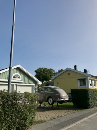 A classic car is elevated on jacks in front of two suburban houses, one painted green and the other yellow, both with enclosed garages. A streetlight and neatly trimmed bushes line the sidewalk, and trees are visible in the background under a clear blue sky.