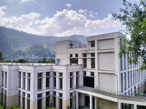 A modern, multistory white building with large windows is set against a backdrop of lush green hills and a partly cloudy sky. Trees are visible in the foreground, framing the building on the right side.