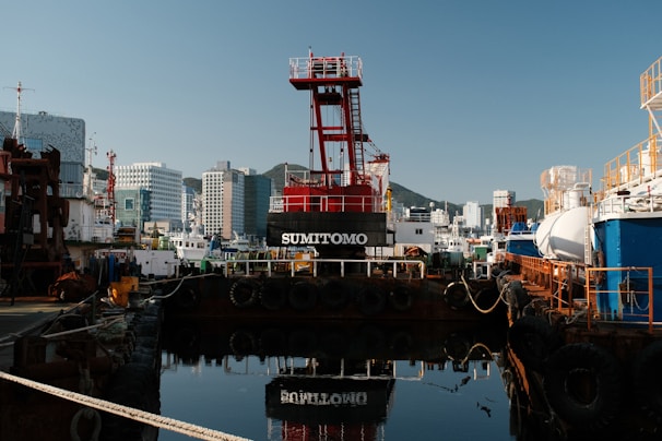 A busy industrial harbor scene with various ships docked alongside large buildings in the background. In the center, a red crane structure with the name 'Sumitomo' on it stands prominent. The reflection of the crane and surrounding ships can be seen in the calm water. The port is surrounded by skyscrapers and hills in the distance.