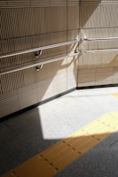 A beige-tiled wall with parallel metal handrails casts a shadow on a light gray tiled floor. A yellow tactile paving strip runs across the floor, providing guidance for visually impaired individuals.