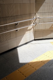 A beige-tiled wall with parallel metal handrails casts a shadow on a light gray tiled floor. A yellow tactile paving strip runs across the floor, providing guidance for visually impaired individuals.