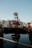 A red and black industrial structure bearing the name 'SUMITOMO' is situated on water. Surrounding it are various other industrial buildings and equipment. The calm water reflects the scene, and tall buildings with mountainous terrain in the background can be seen under a clear blue sky.